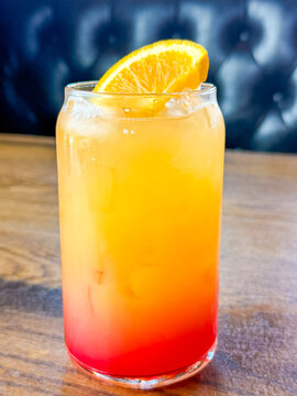 Bright orange and red iced beverage with citrus garnish on a wooden table in front of a dark blue restaurant booth.