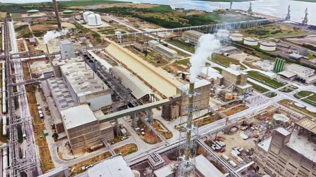 A large industrial area near the shore shows different buildings, storage tanks, and pipes. Smoke rises from a stack while vehicles move around. The site is active and busy during daylight.