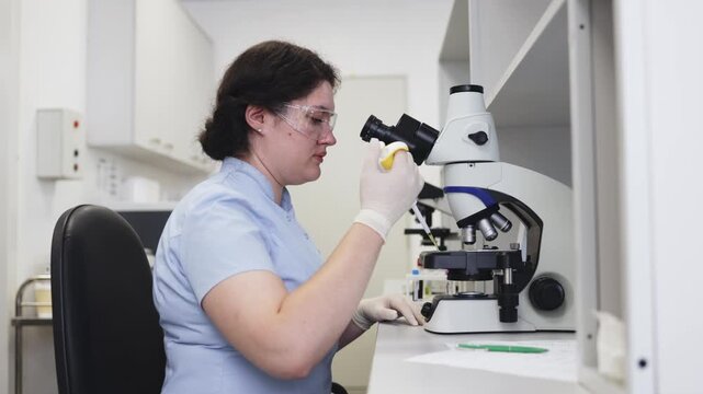 Female laboratory technician wearing safety glasses and gloves applies a liquid biological sample to a microscope slide with a micropipette and examines it under a microscope.