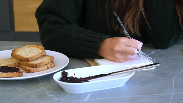 A person is busy writing in a notebook while enjoying a light breakfast of rusks with jam. This scene conveys focus, productivity, and a cozy start to the day.