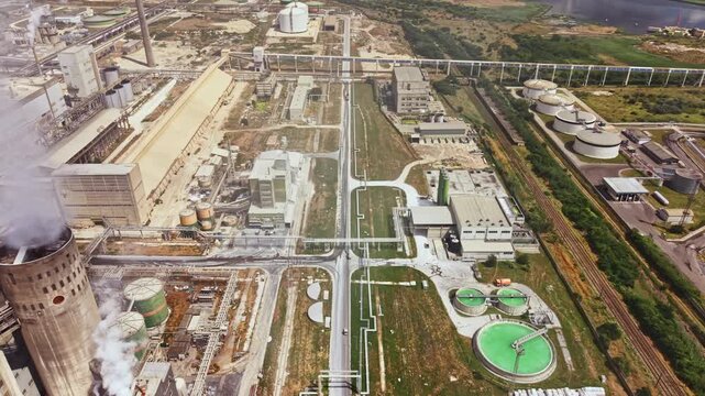 Aerial view of an industrial site with numerous buildings, storage tanks, production areas, and green spaces under clear skies. Pipes connect different sections of the complex.