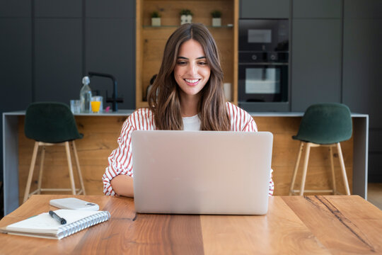 Young woman smiling while working on a laptop at a wooden kitchen table, remote work from home concept, casual home office, notebook and coffee nearby, productivity and focus