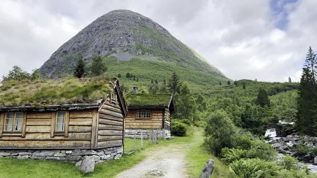 Scenic mountain landscape along the famous Trollstigen road in Norway featuring traditional grass-roof houses, a crystal-clear river flowing through the valley, lush forests, and majestic mountains.