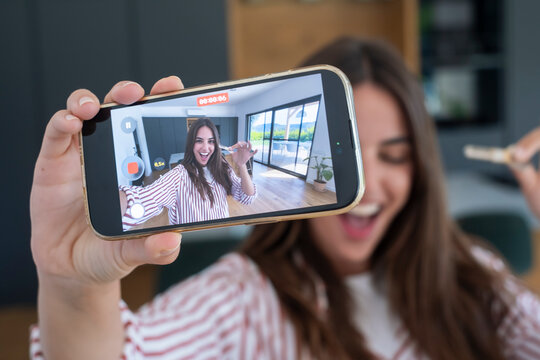 Woman recording a cheerful vlog on a smartphone at home, smiling and holding the phone toward the camera in a bright modern living space