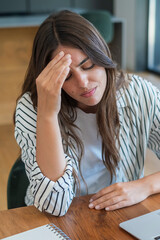Fototapeta premium Young woman with headache touching her forehead while working at a desk, looking stressed and tired during remote work with laptop and notebook