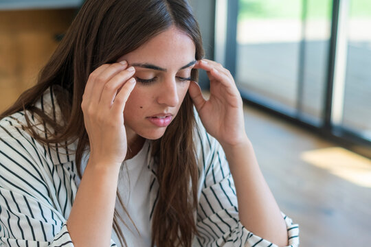 Young woman holding her temples and closing her eyes, looking stressed and tired while studying or working at home near a large window, experiencing headache and mental fatigue