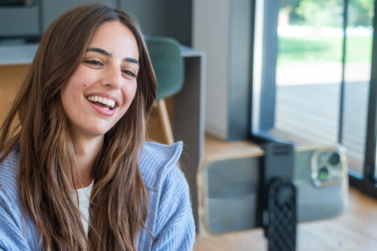Smiling young woman recording a vlog at home with a smartphone on a tripod, casually dressed and engaging with the camera during a relaxed video call or social media content session.