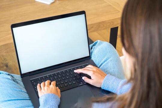 Young woman typing on a laptop while sitting cross-legged on a couch, blank screen visible; casual remote work or studying at home with relaxed, cozy atmosphere
