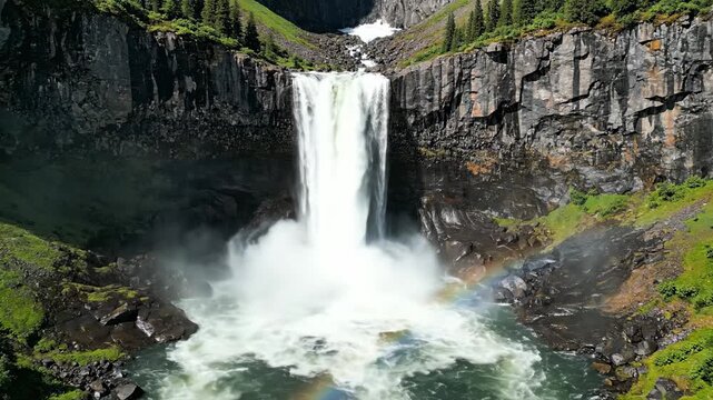 The powerful cascading water of the Hengifoss waterfall crashes into the deep rocky canyon pool while a faint rainbow appears through the misty spray during a bright and sunny summer afternoon
