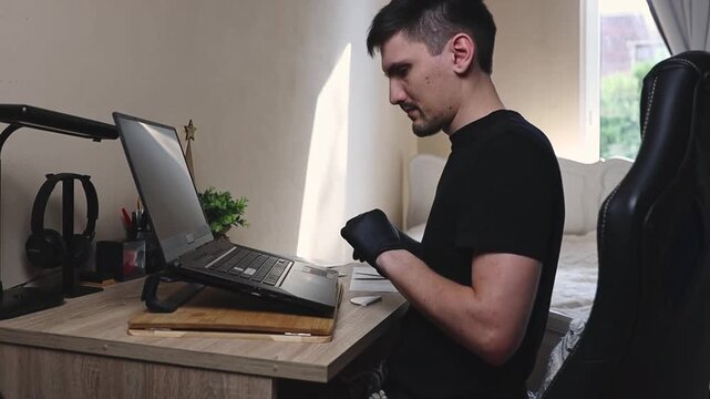 One young handsome Caucasian brunette guy in black latex gloves glues a transparent letter sticker on a laptop keyboard button with special plastic, sitting at the office desk in the bedroom on a summ