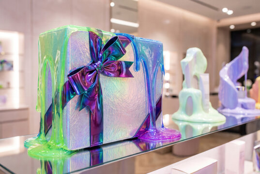 Colorful iridescent gift box with a purple ribbon and melting design displayed on a glass shelf in a modern retail store interior