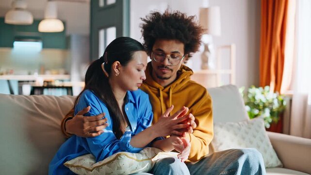 Worried couple sitting sofa holding hands. Multiethnic pair comforting at home.