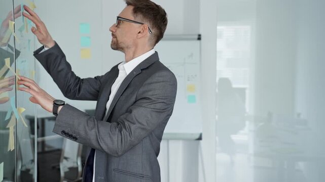 Business professional deeply contemplating project strategy, analyzing ideas and planning solutions on a glass wall with sticky notes in a modern office environment