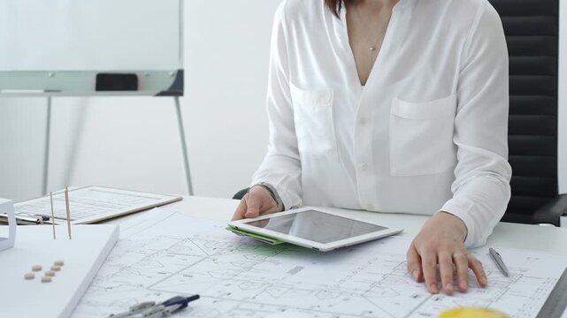 Woman architect analyzing blueprints and architectural models with a digital tablet on a clean desk, working on a construction project in a bright office environment