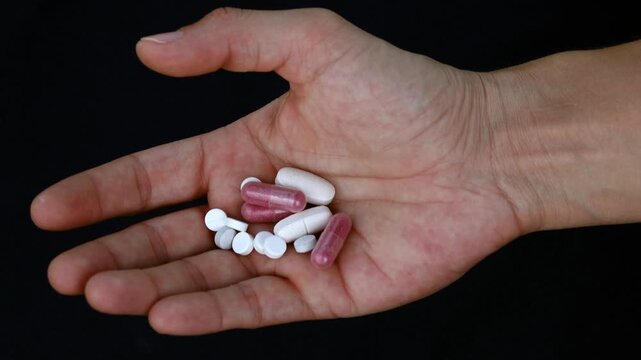 Hand holding various pills and capsules against a dark background. A person's open palm displays a collection of white and pink pharmaceutical capsules and tablets symbolizing medication and treatment