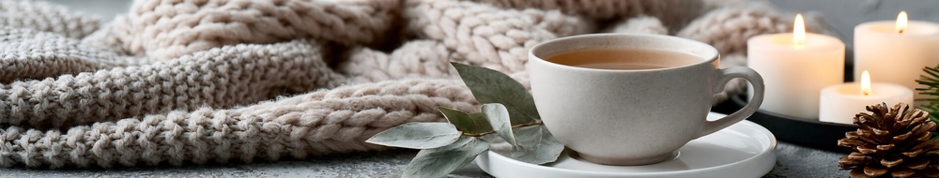Beige knit blanket with ceramic cup of tea, eucalyptus sprig, pine cone and lit candles on a textured tabletop