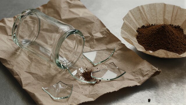 Broken glass jar spills coffee grounds on brown paper, symbolizing loss or destruction.