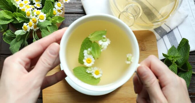Woman with cup of tea, mint and chamomile flowers at wooden table, top view