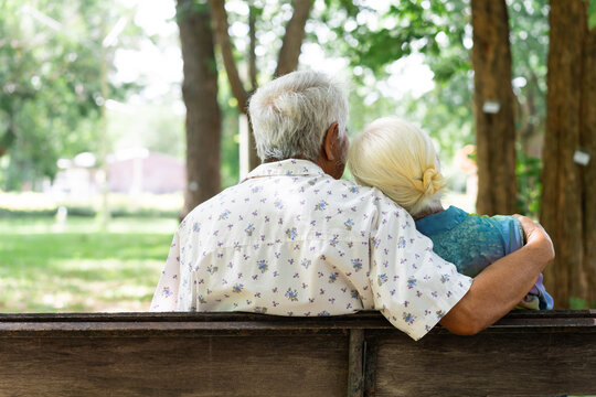 Old couple sits close on park bench. Back view shows quiet, affectionate moment. Tree shadows dappled the grass behind them. Arm around shoulder speaks of comfort and trust