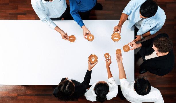 A diverse team of professionals gathers around a white table, collaboratively engaging with gears and cogs, symbolizing teamwork and innovative strategies in a modern workspace. Amity