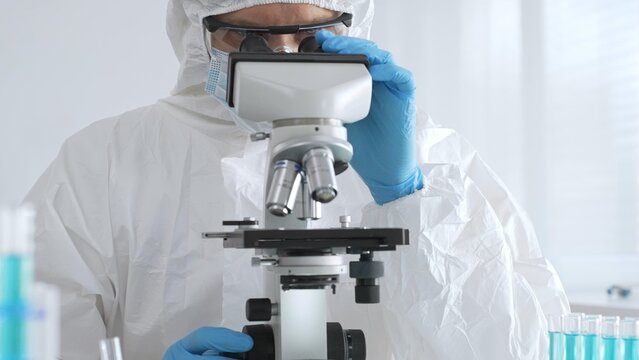 A male doctor in a protective suit and gloves uses a microscope while analyzing samples, conducting research in a laboratory. Close-up of test tubes with blue liquid