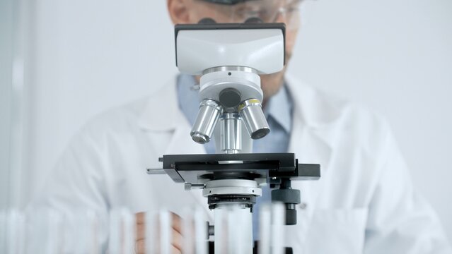 Scientist wearing safety glasses performing laboratory research, looking through an optical microscope, adjusting focus, studying samples in a medical or biotech lab