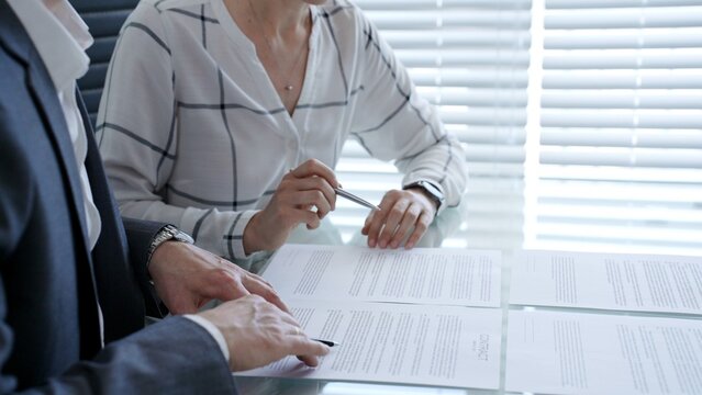 Business professionals reviewing a contract document at a table. Pointing at clauses with a pen. Discussing legal terms and financial agreements during an official meeting