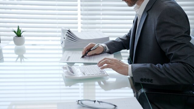 Businessman using a calculator and pen, examining financial documents and reports at a desk, a large stack of papers nearby, representing analysis, accounting, and meticulous planning