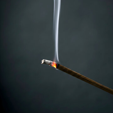 A single thin glowing incense stick burning against a dark solid grey background with rising white smoke