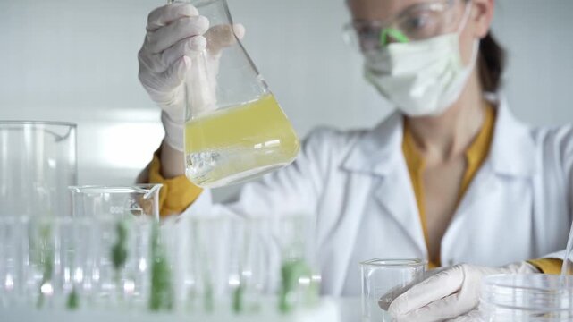 Woman scientist wearing protective gear holding a beaker with yellow liquid while working in a laboratory, close up. Science and medicine