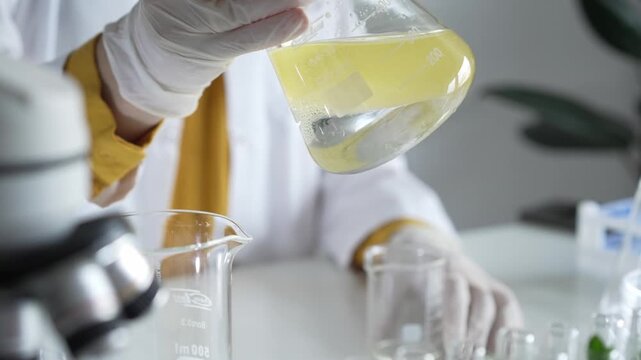 Unknown scientist wearing white protective gloves is carefully shaking up a yellow chemical solution inside an Erlenmeyer flask in a laboratory, close up. Science and medicine