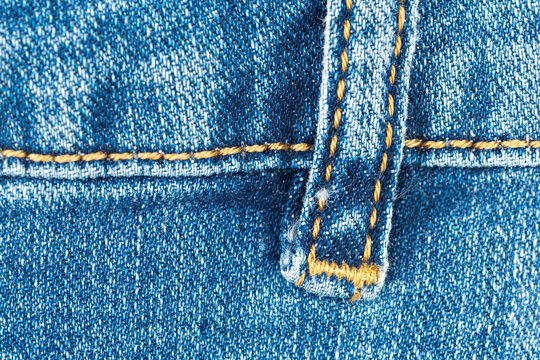 Macro photograph of blue denim fabric. Shows detailed texture of cotton weave, belt loop and prominent horizontal seam with yellow thread. Classic background for fashion or textile concepts