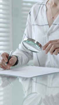 Woman's hands using a magnifying glass to scrutinize a document while holding a pen, researching important information for legal, financial, or academic analysis and validation