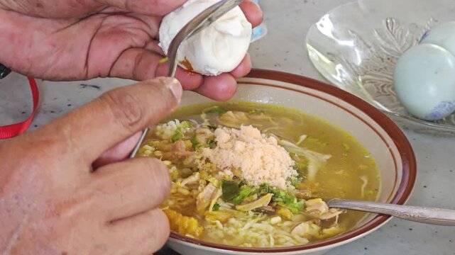 Close up of a person using a spoon to cut and add a salted duck egg into a bowl of traditional Indonesian chicken soup with rice and noodles