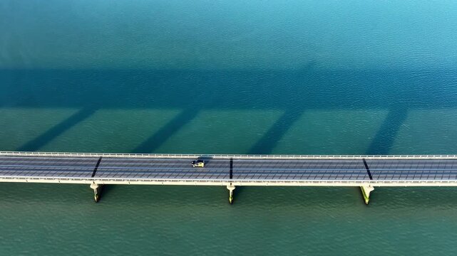 Aerial bridge crossing calm teal water with long diagonal pier shadows and clean geometric lines stretching across wide open landscape, with bridge, aerial, topdown, pier detail