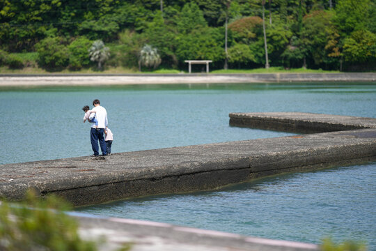 Man holds child on rocky jetty near calm water. Green trees line distant shore behind the pier. Light breeze rustles leaves above the quiet lake. Father and child stand close, sharing quiet moment