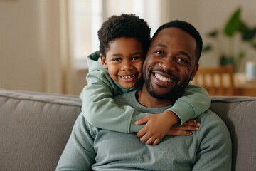 Fototapeta premium Happy African American father, young son embrace on sofa at home. Smiling man with child hug sitting on couch in living room. Carefree boy rests head on dad shoulder. Authentic family connection,