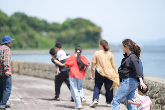 Sunlit seaside path with people walking. Group strolls along stone seawall under clear sky. Woman with child walks beside others casually. Man in hat stands apart, observing the scene