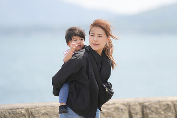 Woman carries child along coastal path. Wind tousles her hair gently. Child smiles, resting close to her chest. Sunlit sea stretches behind stone wall. Perfect for family, travel, or nature themes © Masakazu Tokashiki