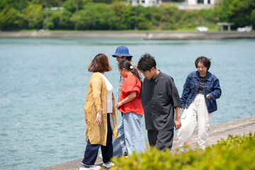 Five friends gather near calm waters. They chat, smile, and enjoy the seaside air. Greenery frames the tranquil lakeside setting. Perfect for social, travel, or lifestyle themes © Masakazu Tokashiki