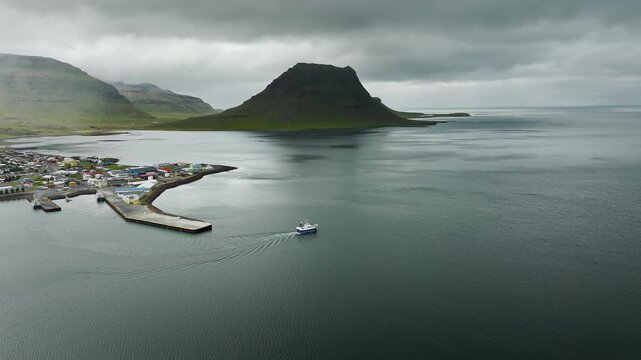 Fishing vessel leaving harbor beneath overcast sky beside coastal village and flat topped mountain with wake tracing movement into open fjord, with boat, wake, harbor, pier detail