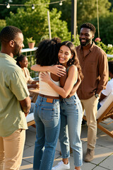 Obraz premium Multiethnic group of friends gather on outdoor terrace at sunset. Two young women hug men smile, talk nearby. People enjoy social time together during summer weekend party. Diverse friends relax at