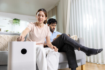 Obraz premium Happy Asian woman adjusting air purifier setting while relaxing on modern living room sofa with man reading book in background breathing clean fresh healthy indoor atmosphere