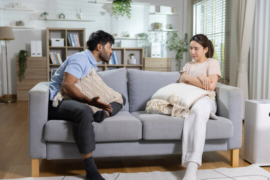 Young Asian couple having serious argument on gray sofa inside bright modern living room feeling frustrated and angry with crossed arms showing emotional tension and relationship conflict
