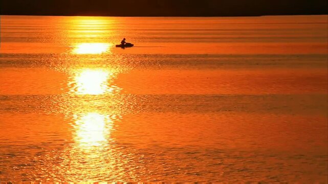 A solitary figure navigates tranquil waters, bathed in the sun's fiery embrace. Golden reflections dance across the surface, painting a breathtaking scene of serene solitude.