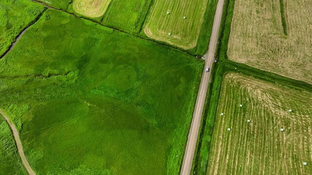 Aerial countryside road with car moving along dirt track between green pasture and harvested fields, with aerial, road, car, farm detail, showing pasture, field, hay, bale context