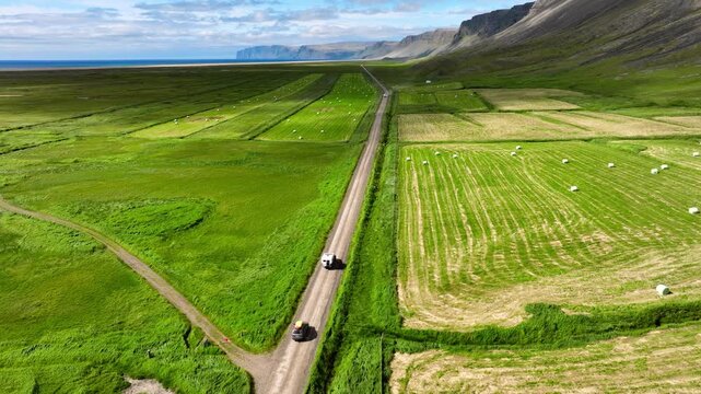 Aerial convoy traveling along straight gravel road through coastal farmland with green fields, rugged cliffs, open ocean horizon, and strong geometric landscape lines