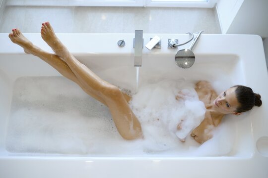 A slender woman enjoys a relaxing bubble bath in a modern bathroom filled with aromatic soap foam. Soft sunlight filters in, adding warmth to the serene atmosphere
