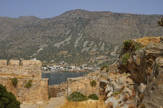 The remains of ancient stone structures in front of a mountainous landscape with a nearby village, Venetian Sea Fortress, Leper Island, Spinalonga, Elounda Bay, Mirabello Bay, East Crete, Crete, Greek Islands, Greece