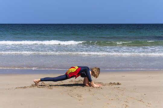 A woman doing yoga on a sandy beach by the sea. Ashva Sancalanasana, hip opening, variation. Treen Cove, Cornwall, England, Great Britain